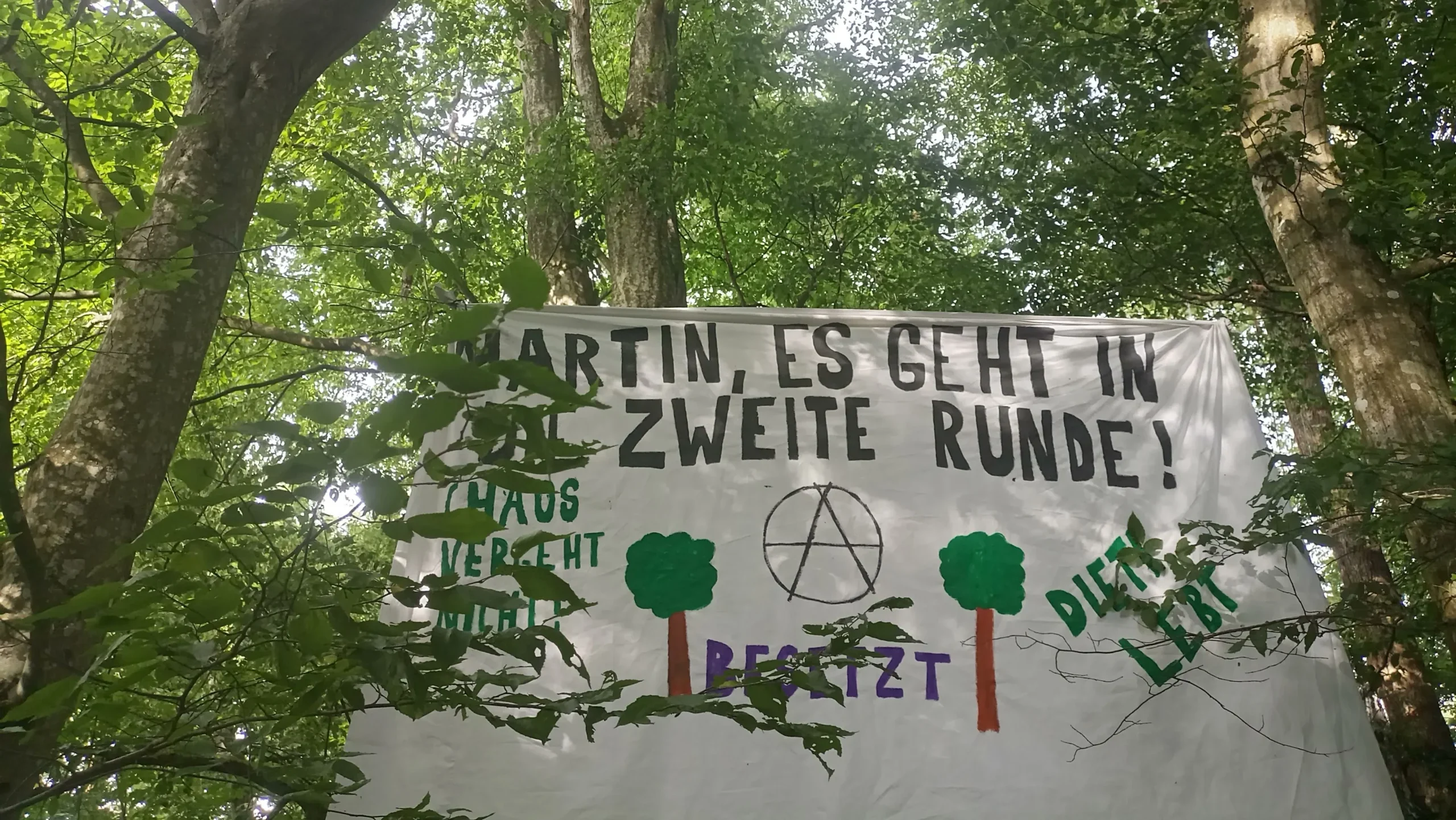 Banner gepannt zwischen Bäumen mit der Aufschrift: Martin, es geht in die zweite Runde. Choas vergeht nicht. Dieti lebt!"